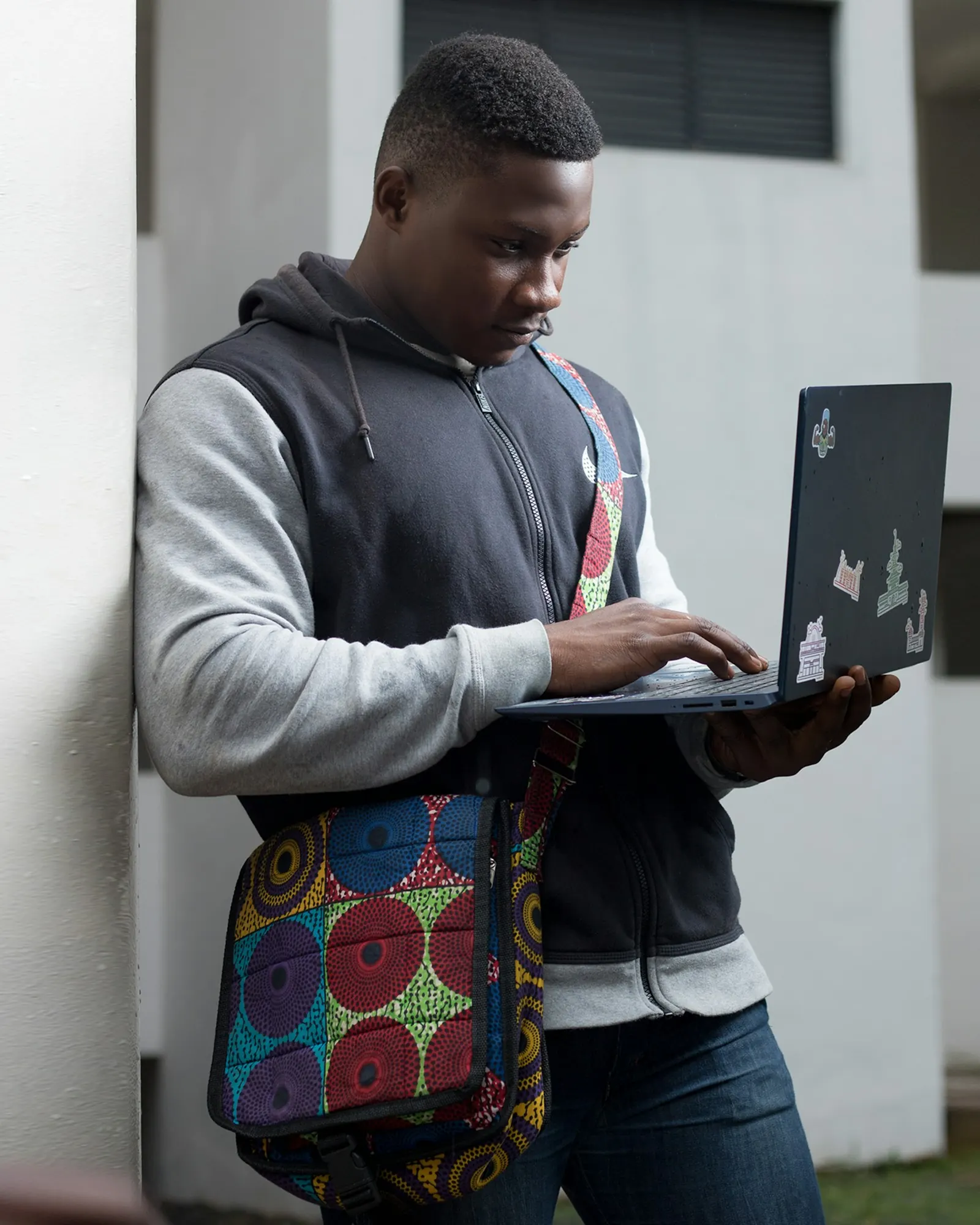 A young man works on a laptop he holds in one arm