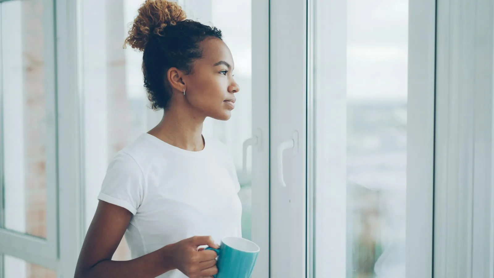 A young woman holds a mug and looks quietly out of a window